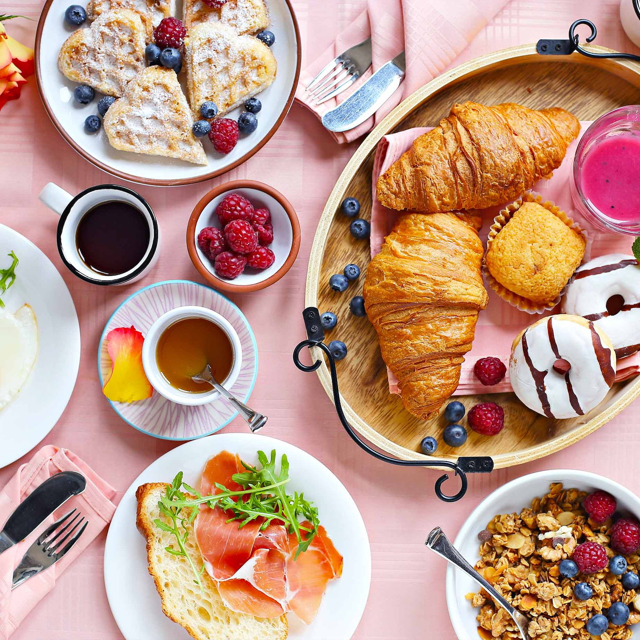 Breakfast flatlay with croissants, berries, toast, coffee and various dishes on pink table