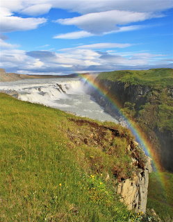 Large waterfall in canyon with rainbow over mist, green landscape and cloudy sky