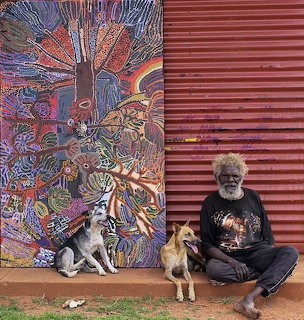 Aboriginal artist Hector Burton sitting with two dogs beside vibrant Indigenous artwork