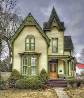 Pale yellow Victorian house with green trim, porch, and tall gables under a cloudy sky