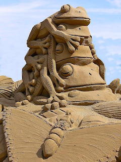 Detailed sand sculpture of a stylized figure on a sandy beach under blue sky