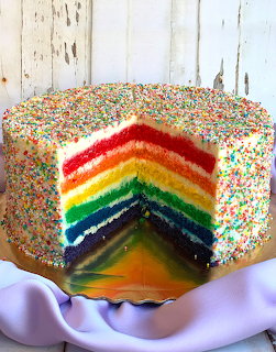 Rainbow layer cake covered in white frosting and colorful sprinkles with a slice cut out revealing six bright colored layers