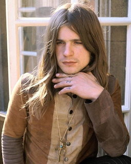 Young man with long brown hair in a brown suede button-up shirt posing near a window