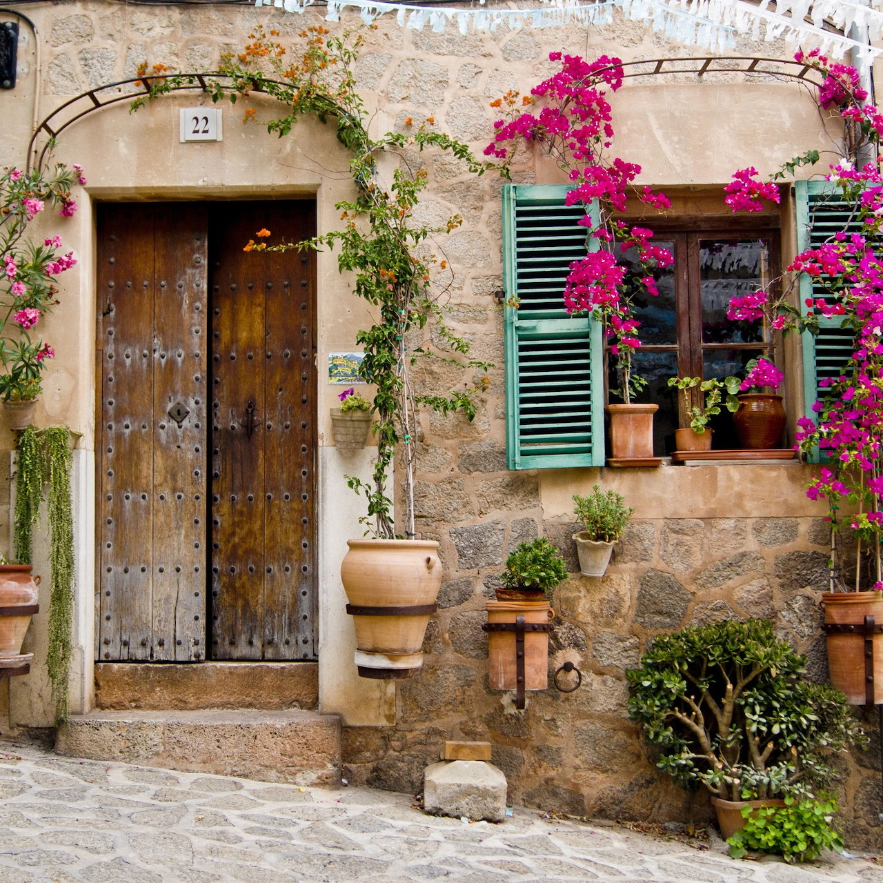 Rustic stone house facade with wooden door, turquoise shutters, pink bougainvillea, and potted plants on cobblestone street