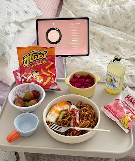 Bed tray with noodles, eggs, strawberries, snacks, drink, and tablet on white bedding