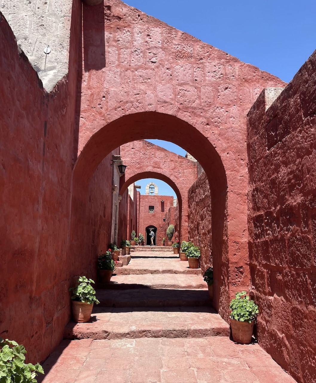 Narrow cobblestone passage through red adobe archways with potted plants and blue sky beyond