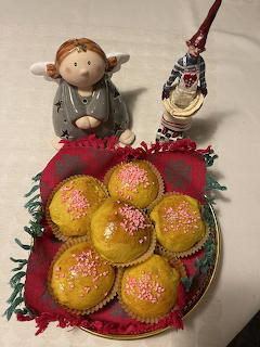Plate of yellow lussebullar with pink sprinkles on a red cloth, with two figurines in the background.