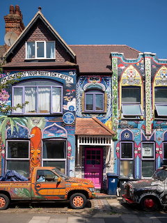 Victorian house exterior painted with bright colorful murals and graffiti art, two decorated cars in front