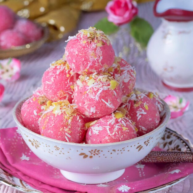Stack of pink coconut-covered truffles with gold flakes in ornate white bowl, pink roses and teapot behind