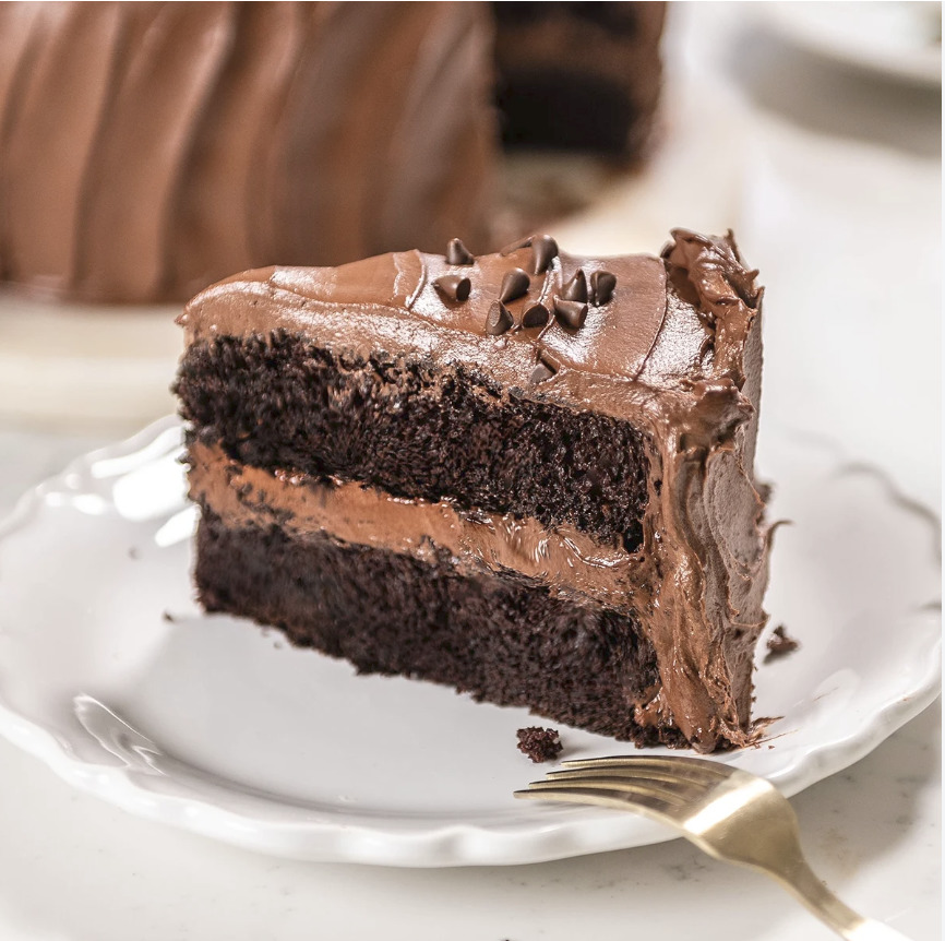 Slice of chocolate layer cake with frosting on a white plate beside a gold fork