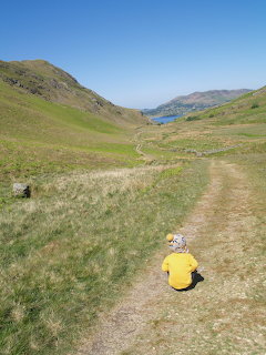 Person in yellow jacket sitting on a dirt path in a green valley with a lake and hills in the background