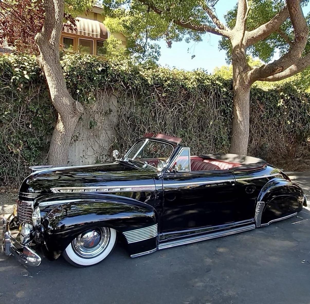 Black vintage convertible parked under trees beside a wall