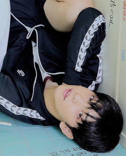 Young man with black hair in a black track jacket reclining against a pale blue tiled wall