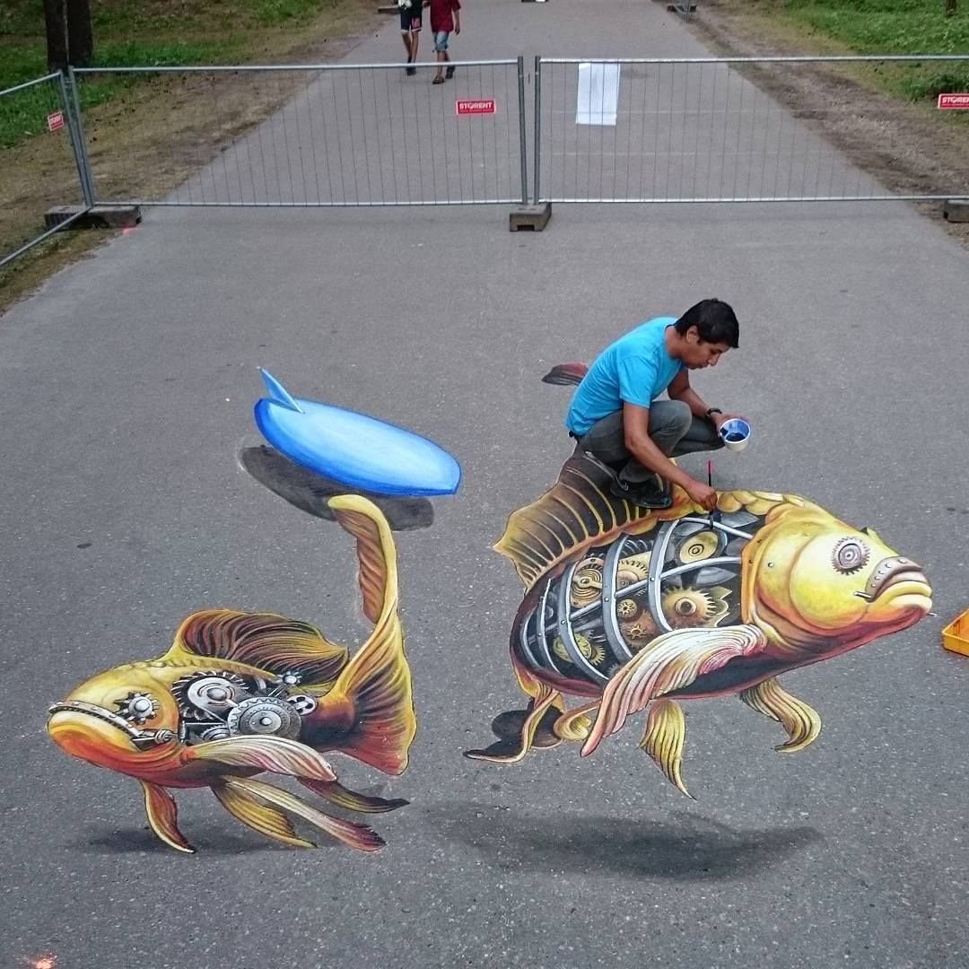Artist drawing 3D chalk art of two koi fish on gray pavement creating illusion of depth