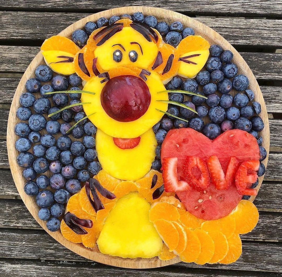 Fruit art on a blueberry-filled plate with a smiling animal figure and a watermelon heart that says LOVE