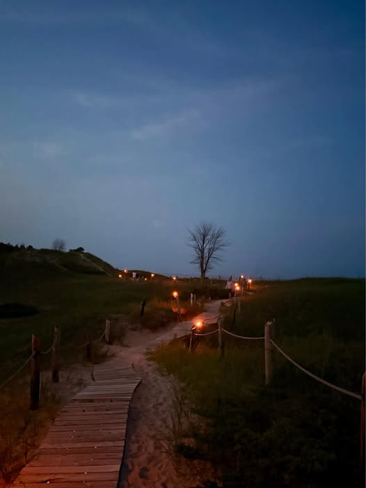 Wooden boardwalk path through sand dunes at dusk, lined with lanterns, lone tree, blue twilight sky