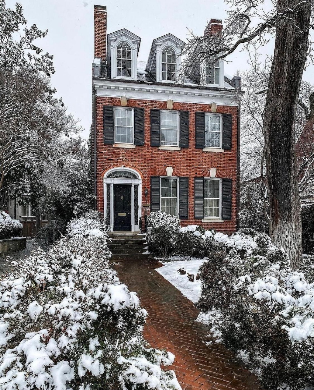Red brick colonial house with black shutters covered in snow, flanked by snow-laden trees and shrubs