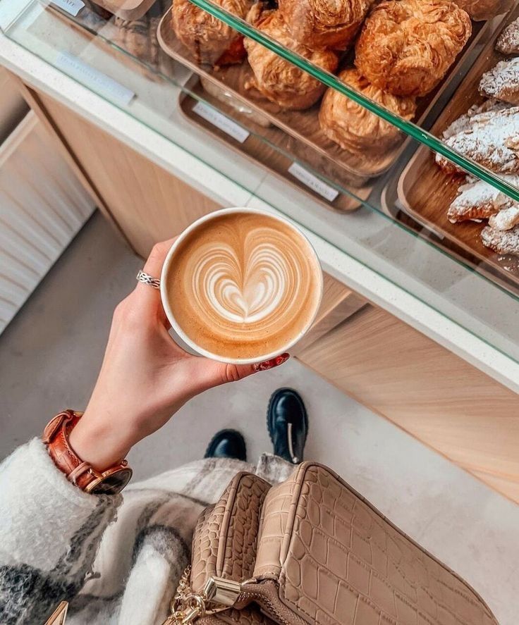 Hand holding latte with heart design over bakery display case filled with croissants and pastries