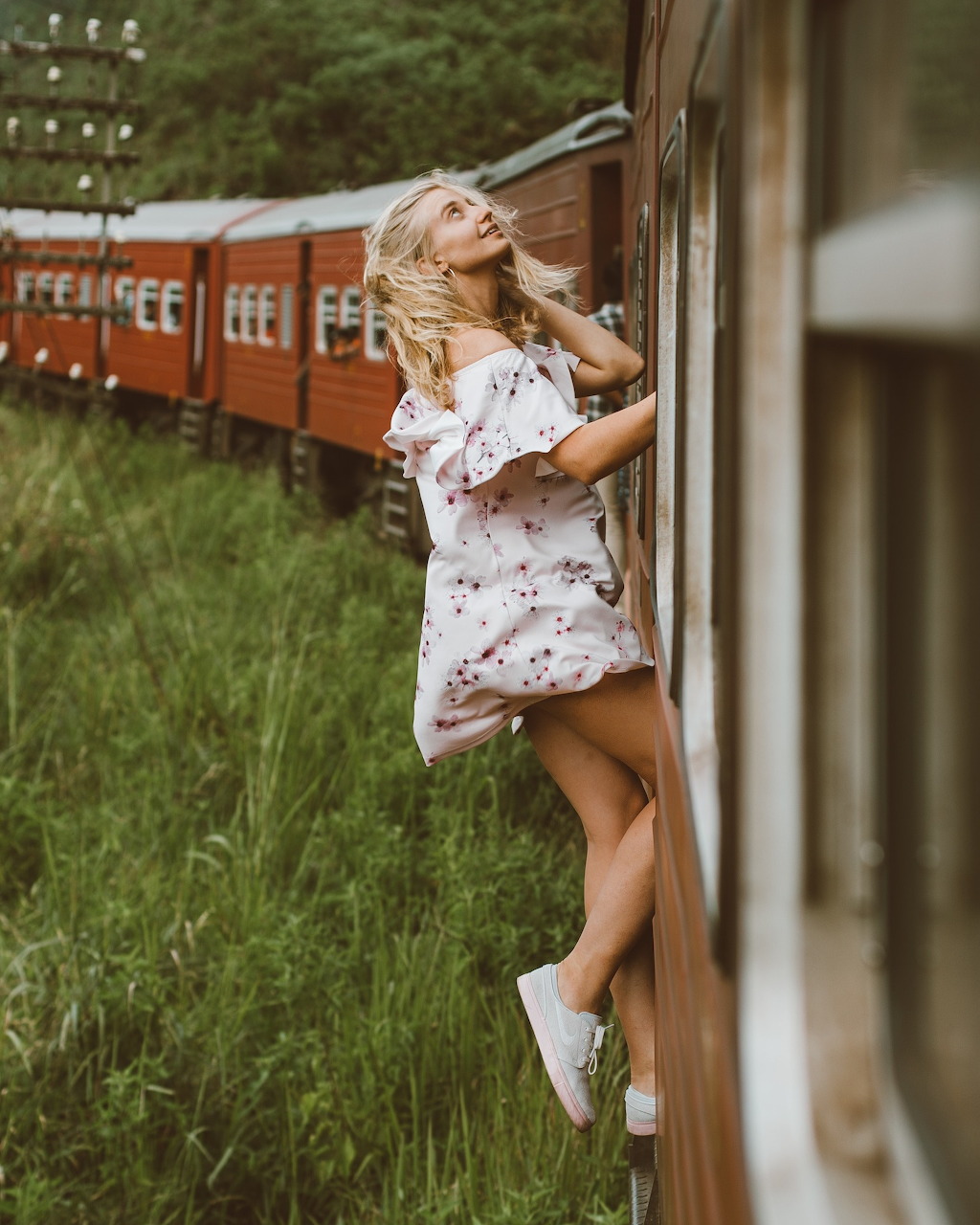 Woman in floral dress and straw hat leaning out of moving train window with countryside behind