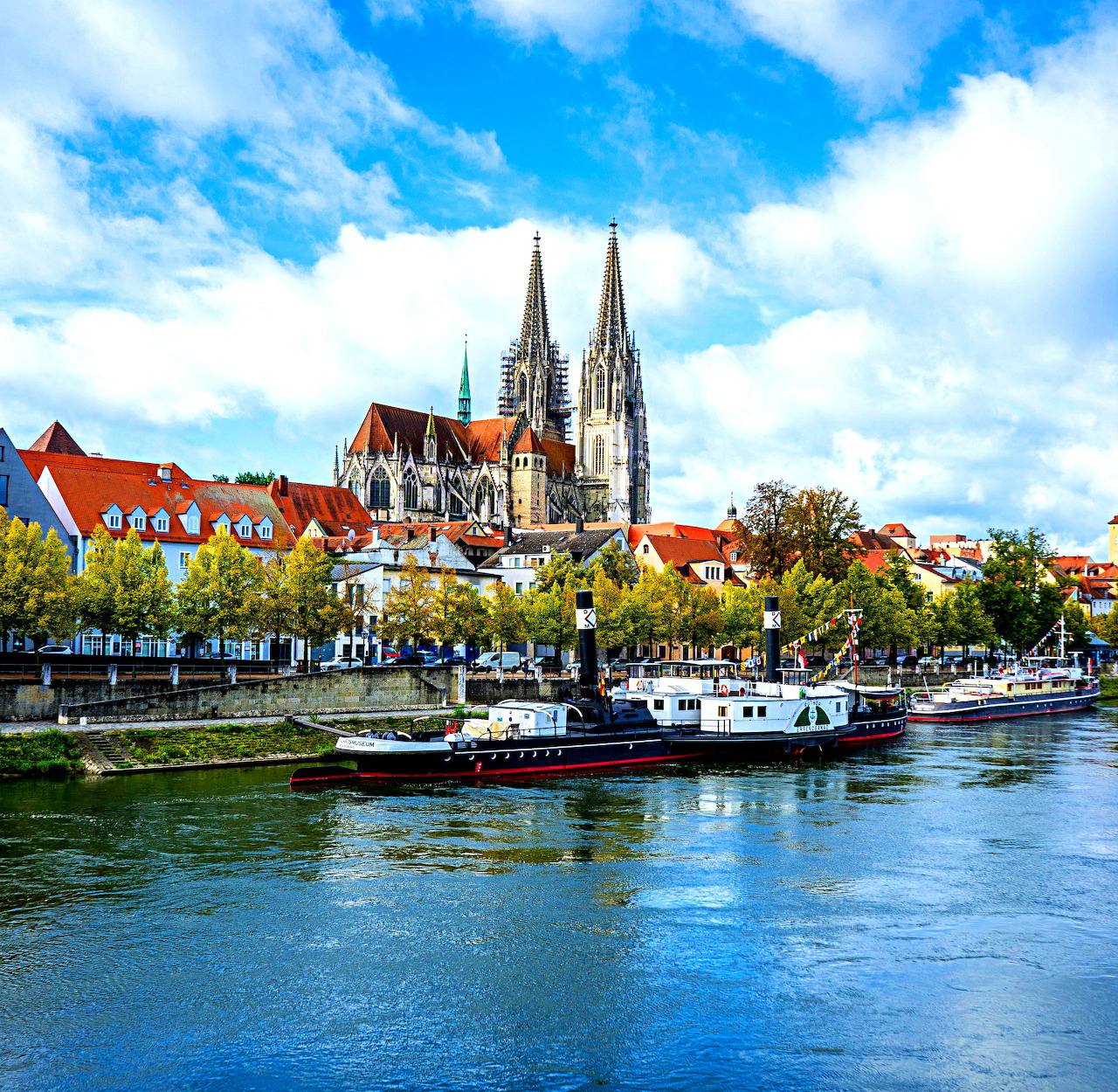 Gothic cathedral with twin spires overlooking colorful riverside buildings and boats on the Danube River in Regensburg