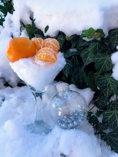 Cocktail glass filled with snow and mandarin oranges beside a glass bauble in a snowy garden with ivy