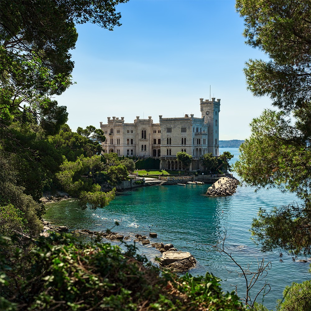 Miramare Castle beside a turquoise bay, framed by trees and a rocky shore.
