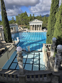 Grand outdoor pool with white Greek temple, marble columns, statues, and bright blue water surrounded by tall cypress trees