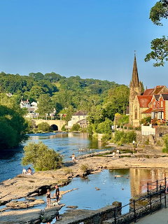 River flowing through a town with a stone bridge, church spire, and green hills in the background