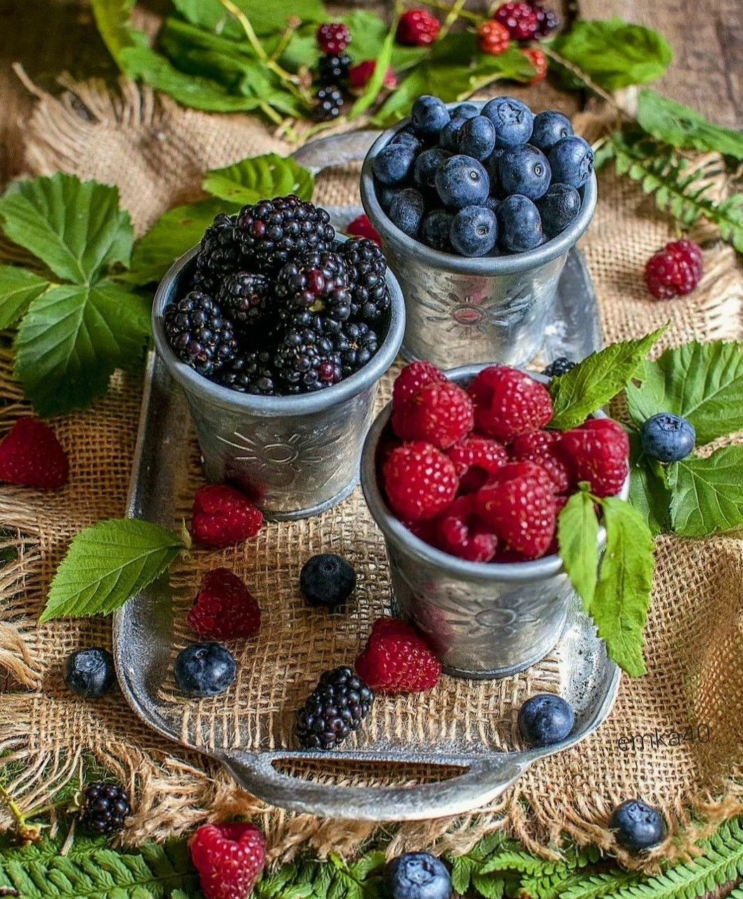 Three metal cups filled with blueberries, blackberries, and raspberries on burlap with green leaves