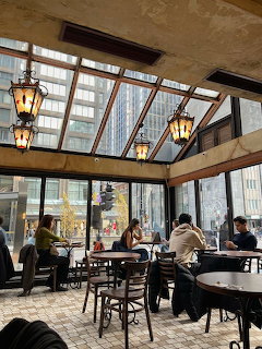 Coffee shop interior with glass roof, hanging lanterns, and patrons at tables with city view through windows