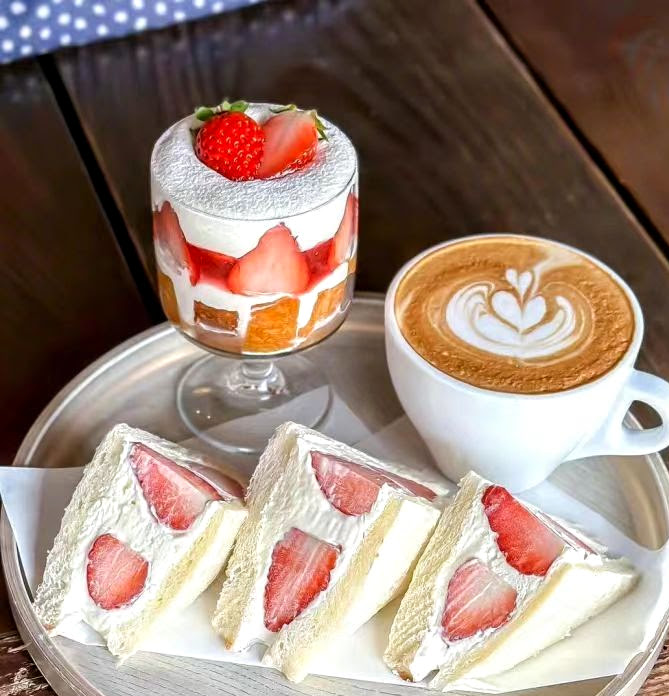 Tray with strawberry cream sandwiches, a strawberry parfait, and a latte on a wooden table