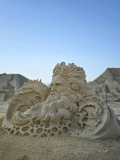 Intricate sand sculpture of a bearded sea god figure with wave details against a blue sky