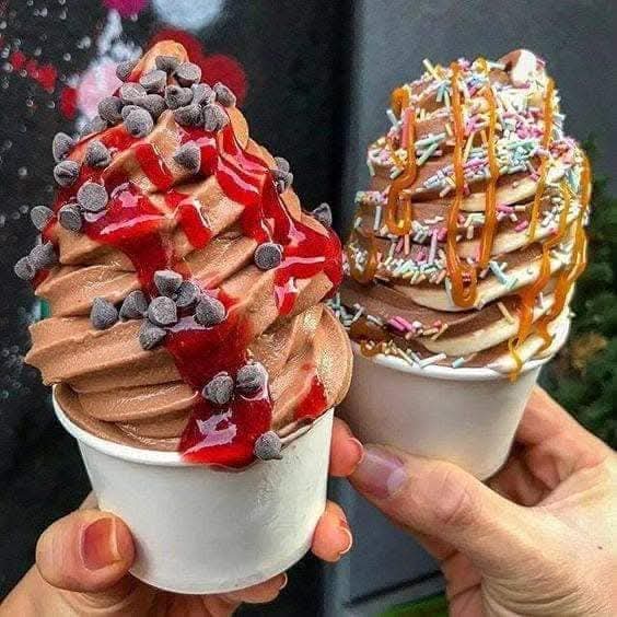 Two hands holding decorated soft-serve ice cream cups with toppings including fruit, wafers, caramel and chocolate