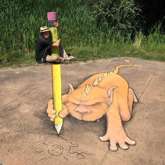3D chalk art of an orange horned monster holding a yellow pencil on pavement with artist crouching nearby