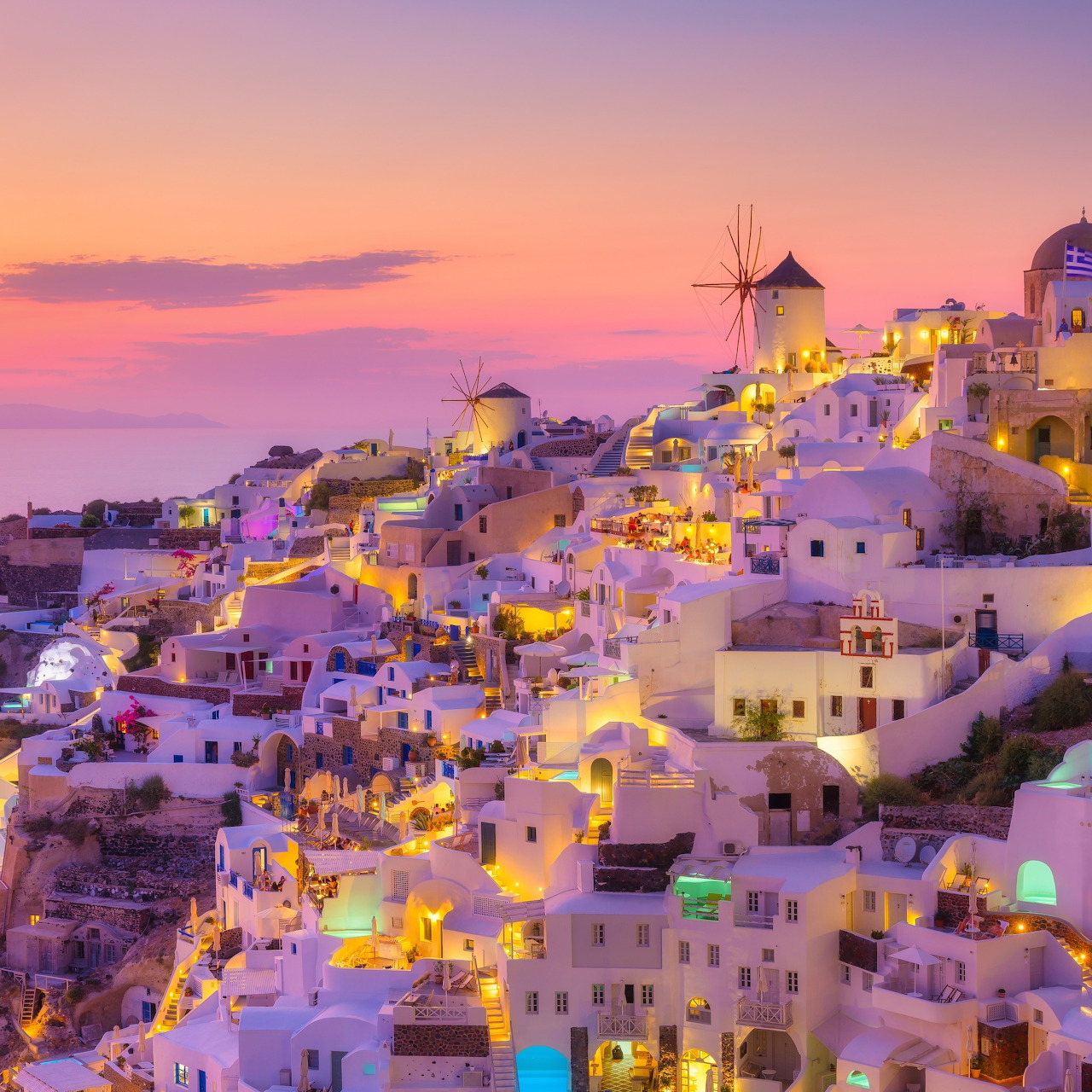 Santorini village at sunset with white buildings, windmills, and glowing lights against a purple-pink sky