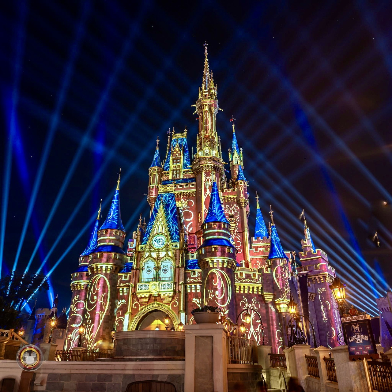 Cinderella Castle lit up at night with colorful projections and blue light beams in the sky