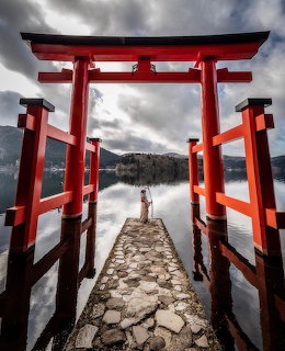 Red torii gate on stone pier over lake with mountains and cloudy sky
