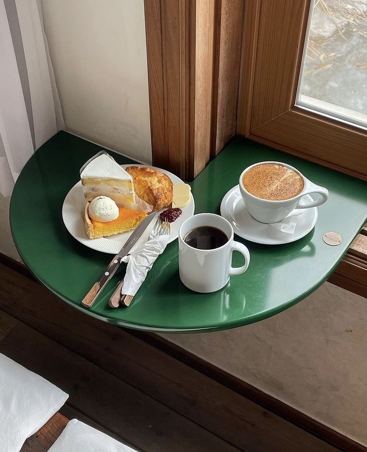 Green half-round cafe table with black coffee, cappuccino, and a slice of cake by a window
