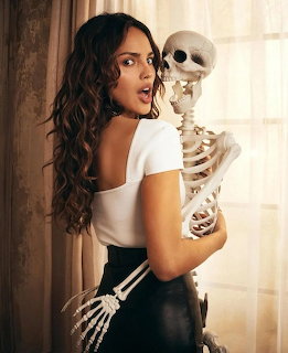 Woman with long wavy hair holding a skeleton prop indoors near a bright window