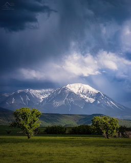 Snow-covered West Spanish Peaks mountain beneath dramatic storm clouds with green meadow and trees in foreground