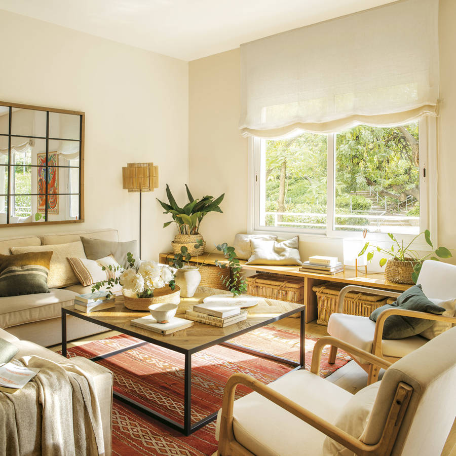 Bright cozy living room with cream chairs, coffee table, large window, plants, and patterned rug