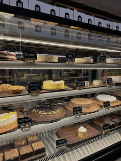 Glass bakery display case with multiple shelves of cakes, chesecakes, and desserts with chalkboard labels