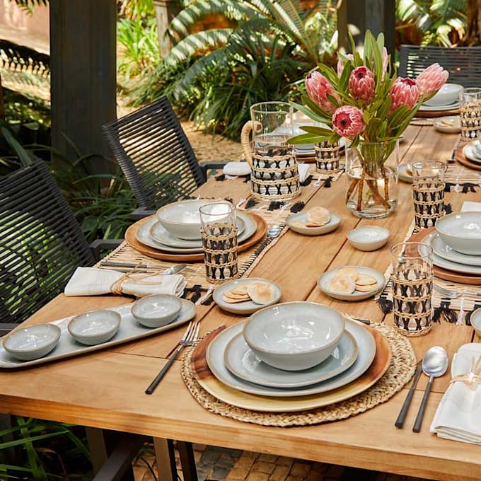 Wooden outdoor dining table set with dishes, patterned cups, and pink flowers in a garden