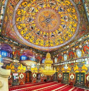 Ornate mosque interior with intricately painted domed ceiling, chandeliers, and red prayer carpet