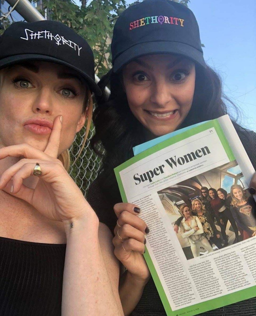 Two women in Shethority baseball caps smiling and holding a Super Women magazine article