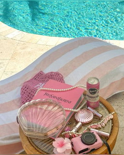 Pink accessories on a striped towel beside a turquoise swimming pool including a shell purse and camera