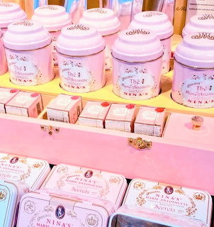 Pink tea tins and small matching boxes arranged on pastel store shelves.