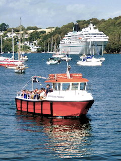 Red passenger ferry on harbor water with sailboats and a large white cruise ship in the background