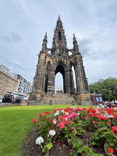 Tall Gothic stone monument with pointed spires and arches surrounded by green lawn and red flowers under cloudy sky