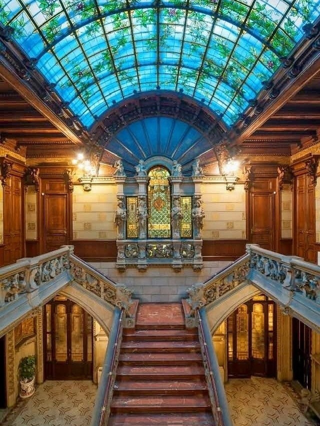 Ornate interior staircase under a blue stained glass ceiling with carved wood and marble railings